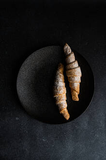 Artistic top view of two sugar-dusted, homemade jam-filled pastries on a dark round plate against a black textured background - ADSF53395