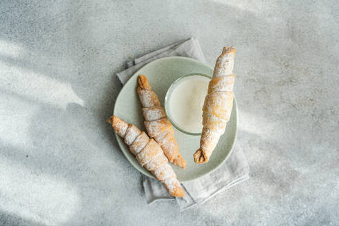 Top view of homemade pastry cones filled with jam and sprinkled with powdered sugar, alongside a glass of milk on a ceramic plate, set on a textured grey surface - ADSF53396
