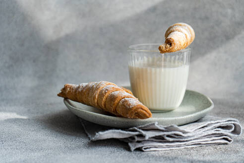 A homemade jam-filled pastry cone dipped in a glass of milk, with another lying on a ceramic plate, against a textured grey backdrop - ADSF53397