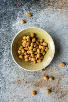 Top view of bowl of salty coated peanuts on a textured surface with some scattered around the bowl - ADSF53398