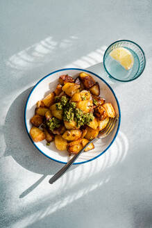 Top view of sunlit roasted potatoes with a dollop of green pesto on a white plate with blue trim, next to a glass of water with lemon, casting a shadow on a textured grey surface - ADSF53401