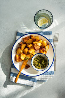 From above of crisp roasted potatoes beside a bowl of green pesto on a blue-trimmed plate, over a blue-striped napkin, with a glass of water with lemon slice in the background, on a textured surface in daylight - ADSF53402