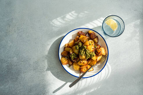 Top view of sunlit roasted potatoes with a dollop of green pesto on a white plate with blue trim, next to a glass of water with lemon, casting a shadow on a textured grey surface - ADSF53403