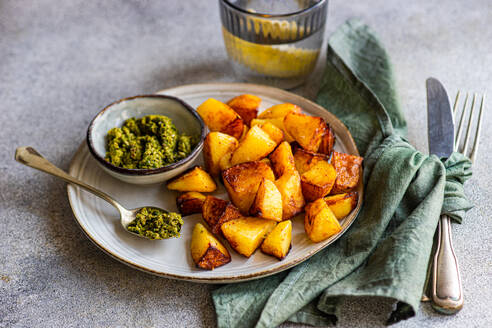 Crispy roasted potatoes on a ceramic plate with a side of green pesto, accompanied by silverware and a green napkin, with a textured grey background - ADSF53405