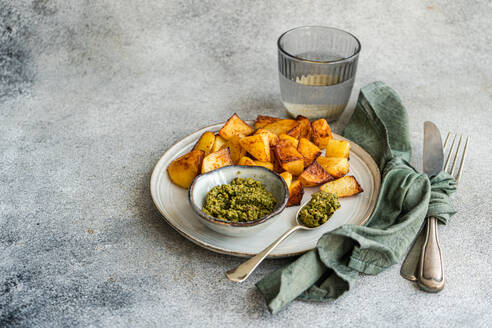 Golden roasted potatoes on a white plate with green pesto in a small bowl, a glass of water, and cutlery wrapped in a green napkin on a textured grey background - ADSF53406