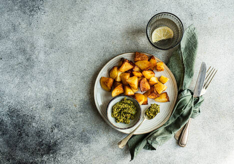 Top view of golden roasted potatoes on a white plate with green pesto in a small bowl, a glass of water, and cutlery wrapped in a green napkin on a textured grey background - ADSF53407