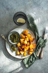 Top view of golden roasted potatoes on a white plate with green pesto in a small bowl, a glass of water, and cutlery wrapped in a green napkin on a textured grey background - ADSF53408