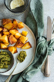 Close-up of crispy roasted potatoes with green pesto, cutlery, and green napkin on a plate, with a glass of water and lemon in the backdrop on a speckled grey surface - ADSF53409