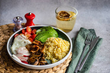 A nutritious meal consisting of cooked bulgur wheat, a sunny side up egg, sliced avocado, fresh cherry tomatoes, radishes, and sautéed mushrooms on a woven placemat with cutlery and a glass of lemon water on the side - ADSF53410