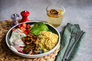 A nutritious meal consisting of cooked bulgur wheat, a sunny side up egg, sliced avocado, fresh cherry tomatoes, radishes, and sautéed mushrooms on a woven placemat with cutlery and a glass of lemon water on the side - ADSF53410