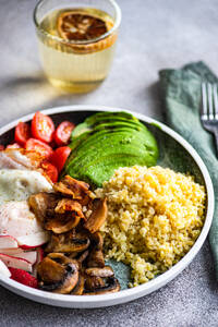 Close-up of a balanced lunch with a portion of bulgur wheat, a fried egg, avocado slices, cherry tomatoes, radishes, and brown mushrooms, accompanied by a glass of lemon water - ADSF53411