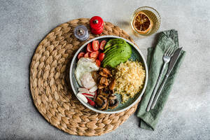 Top view of wholesome plate of bulgur wheat, sunny side up egg, avocado, tomatoes, mushrooms, and radishes, with condiments and a glass of lemon water on a textured background - ADSF53413