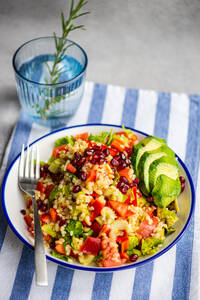 A colorful plate of bulgur salad with pomegranate seeds, fresh vegetables, and avocado slices, presented on a blue-striped linen, with a glass of water and rosemary sprig - ADSF53414