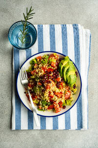 Top view of colorful plate of bulgur salad with pomegranate seeds, fresh vegetables, and avocado slices, presented on a blue-striped linen, with a glass of water and rosemary sprig - ADSF53415