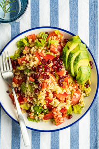 Top view of vibrant plate of bulgur salad with avocado, pomegranate, and diced vegetables on a blue and white striped cloth - ADSF53416