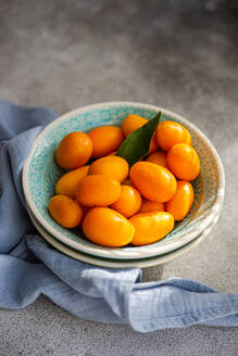 Top view of bowl of vibrant, fresh kumquats with a green leaf, placed on a textured surface with a blue napkin - ADSF53417