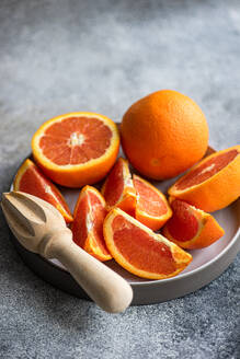 A plate full of ripe, freshly sliced oranges next to a wooden citrus juicer on a textured grey background. - ADSF53419