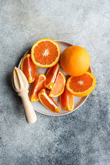 From above sliced and whole oranges arranged on a ceramic plate next to a wooden citrus juicer on a textured gray surface. - ADSF53421