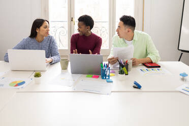 Three professionals actively engaged in a team discussion over documents at a workstation with laptops, colorful stationery, and data charts. - ADSF53596