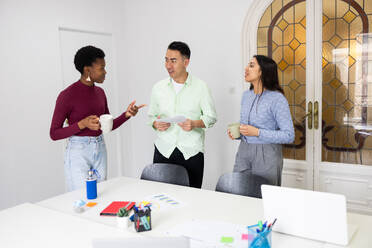 Three diverse professionals engaging in a lively discussion over a project document in a bright office setting, with cups of coffee in hand. - ADSF53598