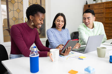 Three colleagues with diverse ethnic backgrounds are sharing a light-hearted moment while collaborating around a table in an office environment, with digital tablets and laptops in use, suggesting a tech-savvy and friendly workspace. - ADSF53607