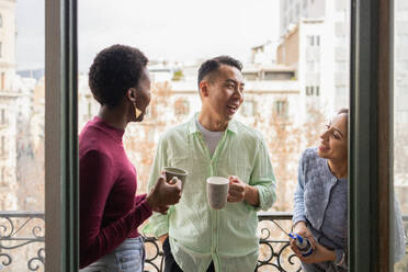 Three friends enjoy a light-hearted moment with mugs of coffee on an urban balcony, sharing laughter and conversation. - ADSF53619