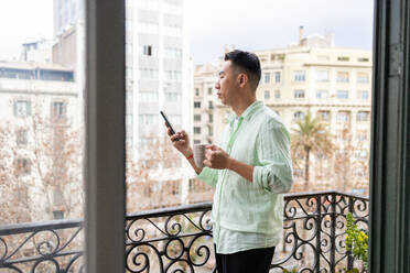 A young asian man holding a coffee mug stands on a balcony with his phone, overlooking a cityscape in a relaxed stance. - ADSF53621