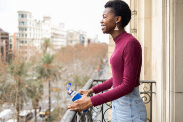 A happy black woman stands on a balcony with a reusable water bottle, overlooking a cityscape, promoting eco-friendly hydration. - ADSF53622