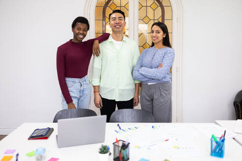 A multi-ethnic group of three professionals stands confidently in a modern office setting, smiling at the camera, with a laptop and work materials on the table in front of them. - ADSF53630