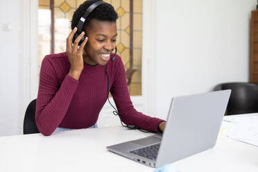 African American woman engaging in a cheerful conversation using headphones with a microphone, seated in front of a laptop, possibly in a customer service or remote work scenario. - ADSF53631