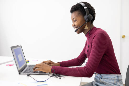 A cheerful customer service representative african female wearing a headset smiles while typing on a laptop at a modern office desk. - ADSF53632