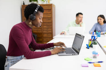 Young professional black woman working at her laptop with headphones in a modern office environment. - ADSF53634