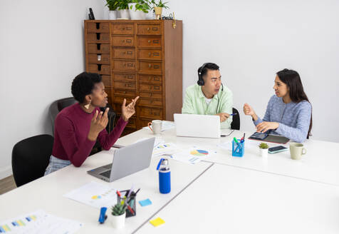 Three professionals engage in a constructive discussion at a white table in a well-lit office space, surrounded by technology and business reports. - ADSF53638