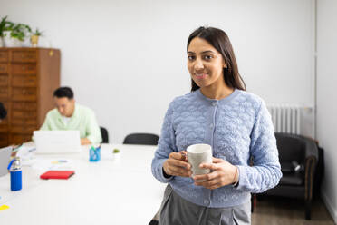 A cheerful professional woman holding a cup of coffee, with a male colleague working in the background at an office setup. - ADSF53639