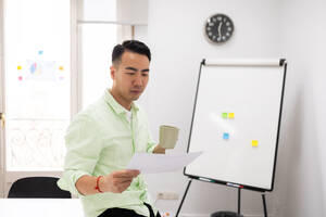 A business professional in a light green shirt holds a coffee cup and reads a document, standing next to a whiteboard in a bright office setting. - ADSF53640