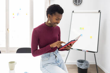A cheerful African-American woman is engaged with a digital tablet, standing in a bright office environment with a clear whiteboard and clock in the background. - ADSF53643