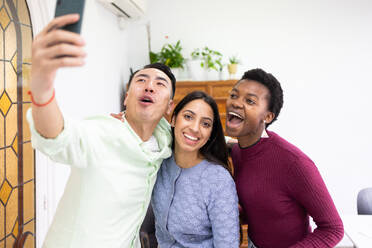 Three cheerful friends from diverse backgrounds share a moment of joy as they pose for a selfie with a smartphone in a bright indoor setting. - ADSF53645