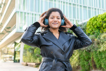 Smiling woman in a trench coat enjoys music on wireless headphones with a modern building and greenery in the background - ADSF53650