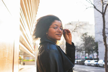 Stylish woman with glasses stands on a city sidewalk at dusk, with the warm glow of the setting sun illuminating her face - ADSF53653