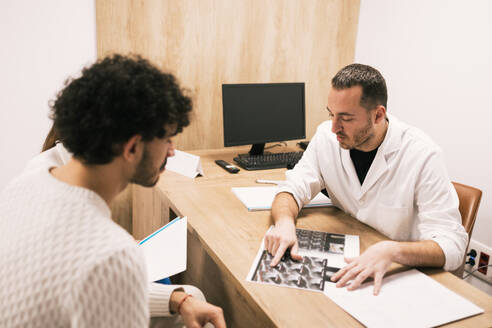 A medical professional examines an X-ray film while discussing the findings with a focused patient during a consultation in a clinic office. - ADSF53660
