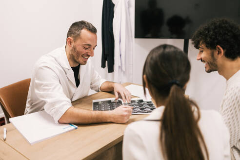 A cheerful male doctor is explaining X-ray results to a young couple in a medical office setting, with a focus on patient care and communication. - ADSF53663