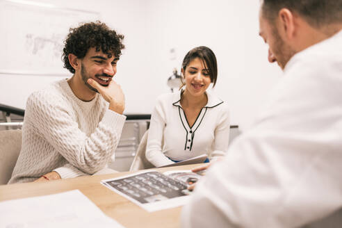 A cheerful male doctor is explaining X-ray results to a young couple in a medical office setting, with a focus on patient care and communication. - ADSF53664