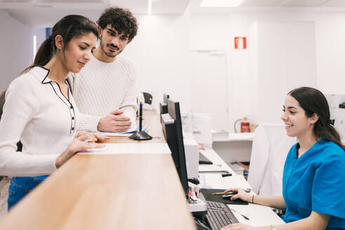 A friendly receptionist assists a couple at a medical clinic's front desk, providing information and guidance with a smile. - ADSF53665