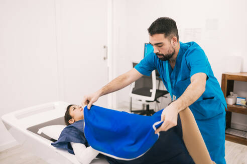 A healthcare professional in scrubs helps a female patient onto an MRI machine, preparing for diagnostic imaging in a clinical setting. - ADSF53667