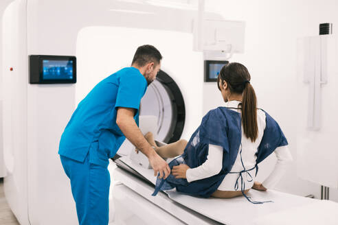 A healthcare professional in scrubs helps a female patient onto an MRI machine, preparing for diagnostic imaging in a clinical setting. - ADSF53668