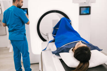Ein Medizintechniker bedient aufmerksam einen CT-Scanner, während ein liegender Patient mit einer blauen Decke zugedeckt auf die Untersuchung wartet. - ADSF53670
