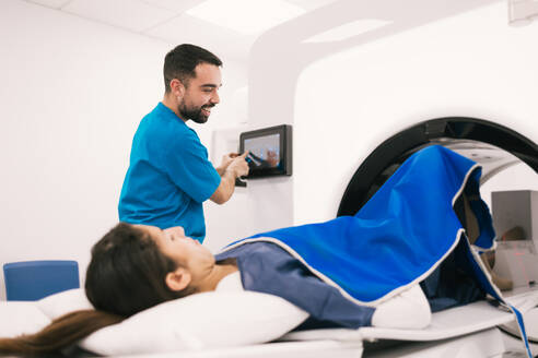 Smiling male radiologist operating a computer while preparing a female patient for a computed tomography scan in a modern medical facility. - ADSF53672