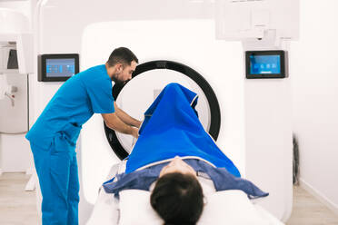 Smiling male radiologist operating a computer while preparing a female patient for a computed tomography scan in a modern medical facility. - ADSF53673