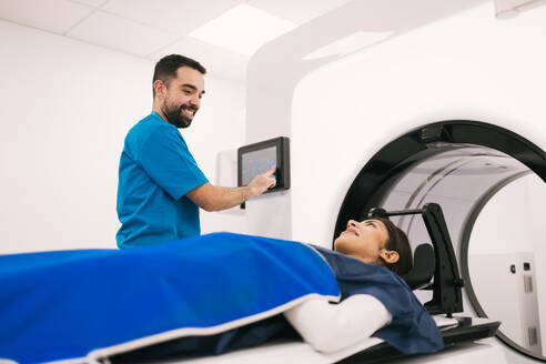 Smiling male radiologist operating a computer while preparing a female patient for a computed tomography scan in a modern medical facility. - ADSF53677