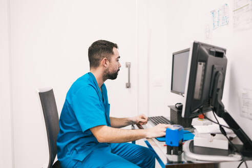 A dedicated male nurse in scrubs is focused on updating patient records on a computer in a medical clinic. - ADSF53680